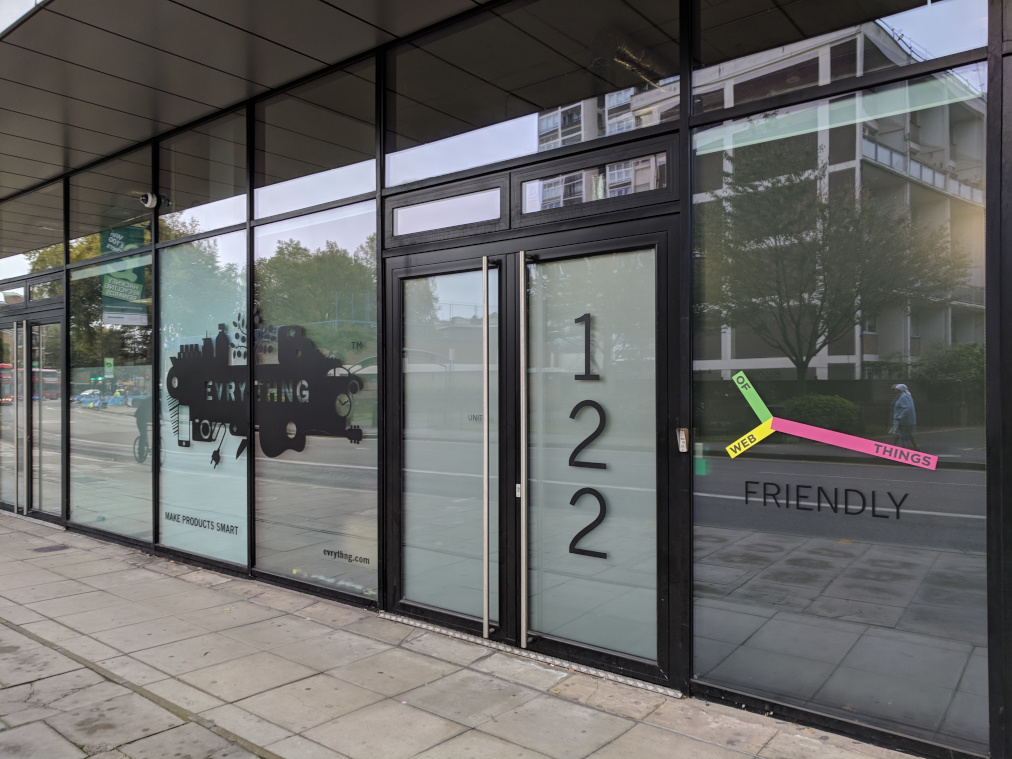 A photograph of the front of an office building with glass doors, the EVRYTHNG company logo and Web of Things community logo.